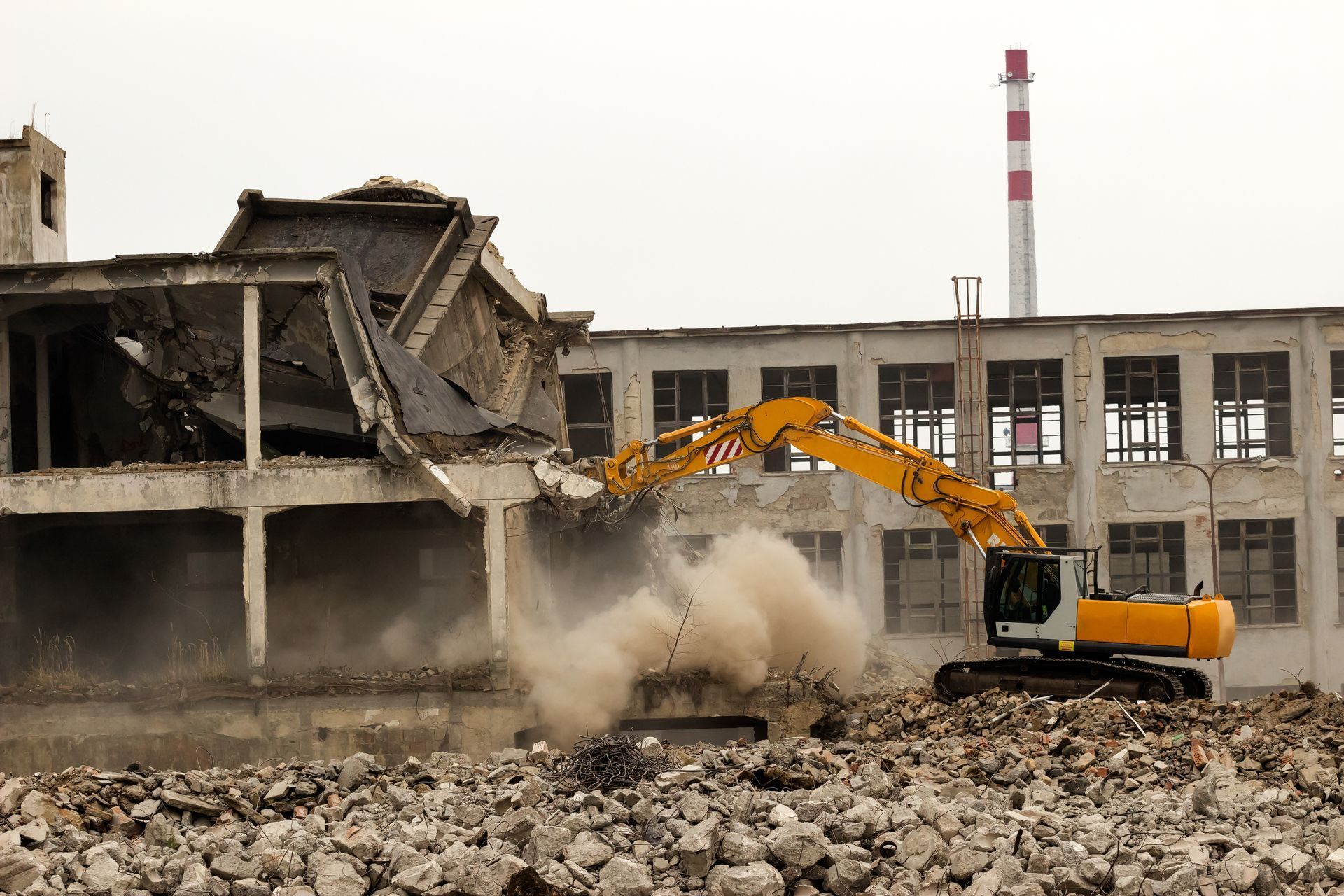 A yellow excavator demolishes a concrete building, kicking up dust; a smokestack is in the background.