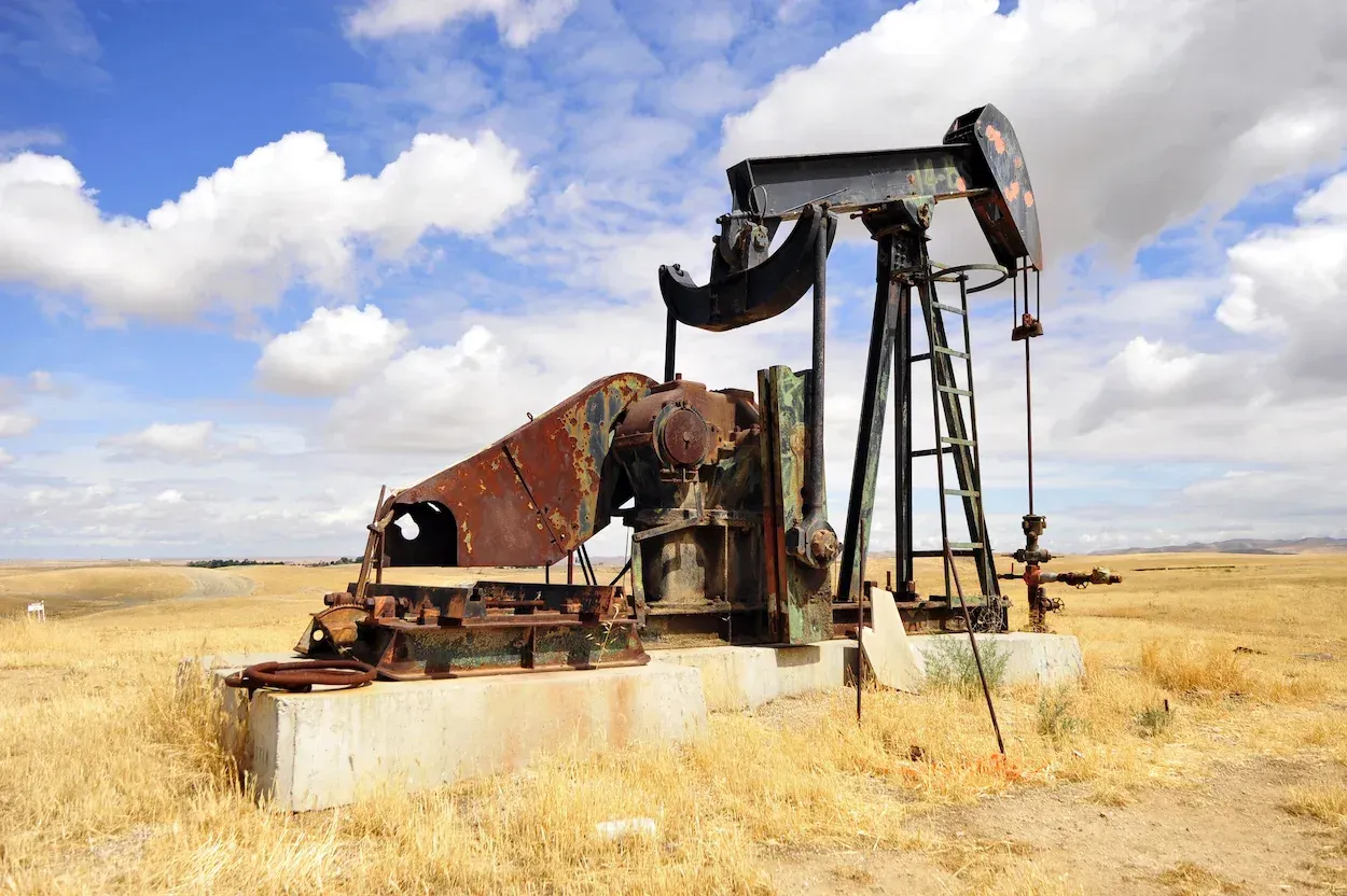 Rusty oil pump in a dry, grassy field under a cloudy blue sky.