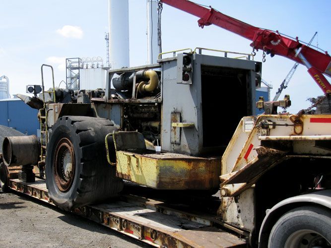 A large, rusted industrial machine on a flatbed trailer, with a red crane arm in the background.