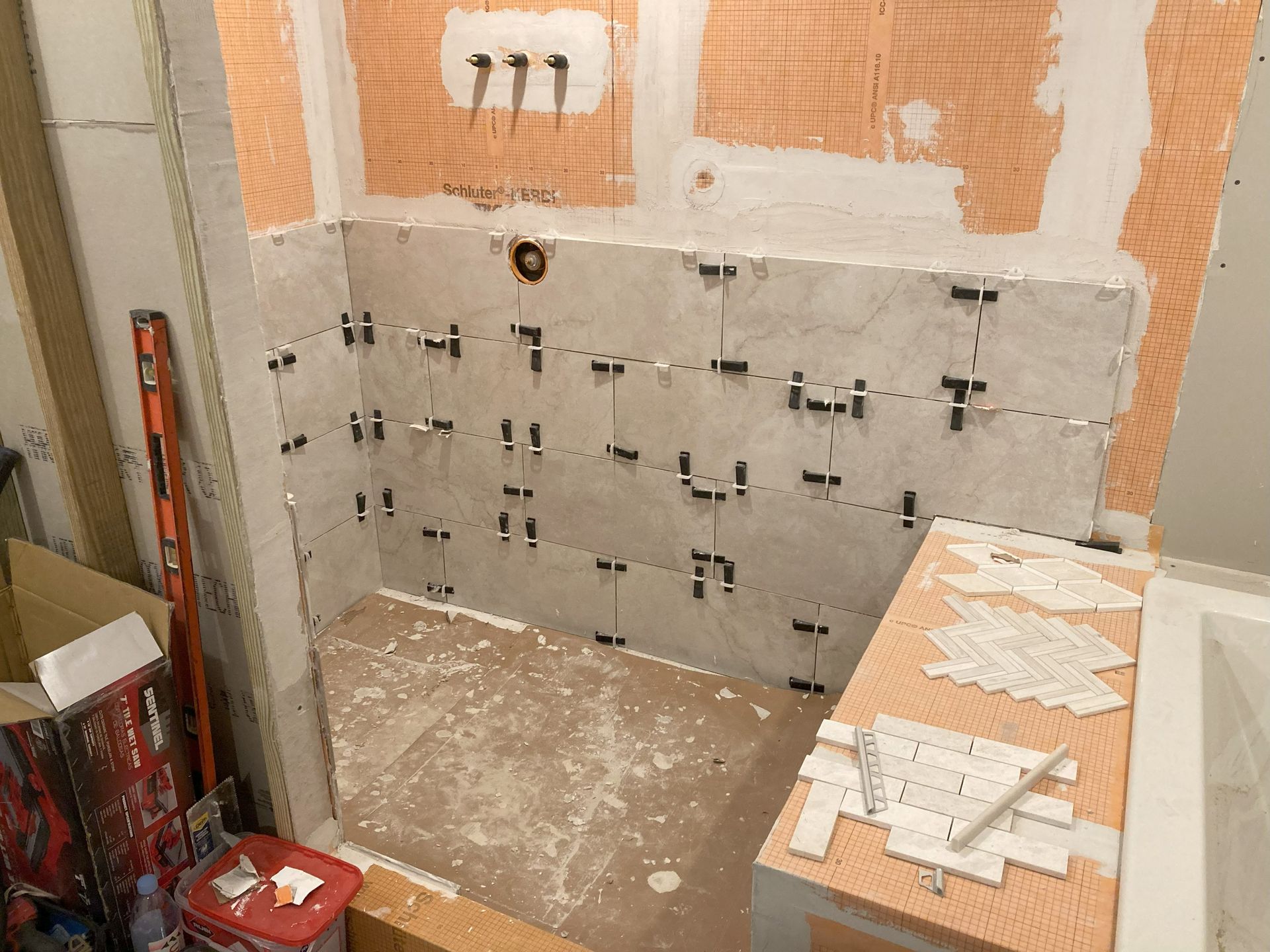 Bathroom under construction; gray tiles installed with spacers on orange waterproof backing, a level, and tile fragments.