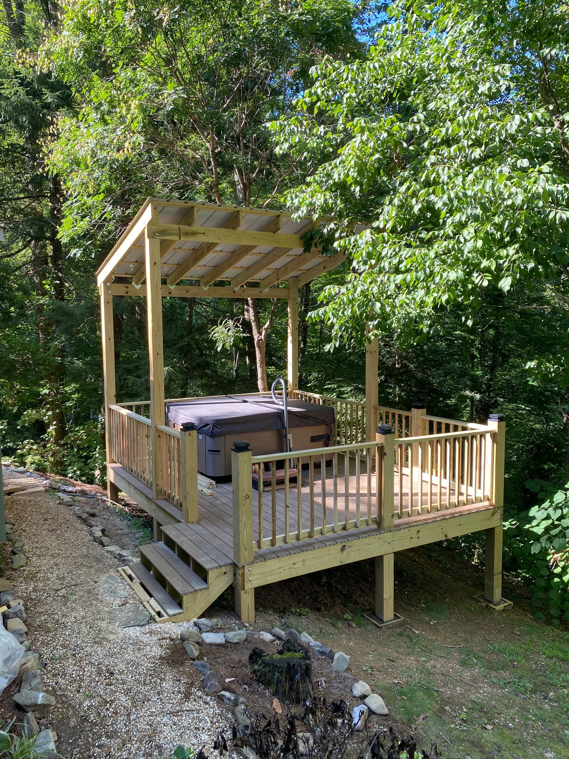 Wooden deck with hot tub, surrounded by railing and pergola roof, in a wooded area.