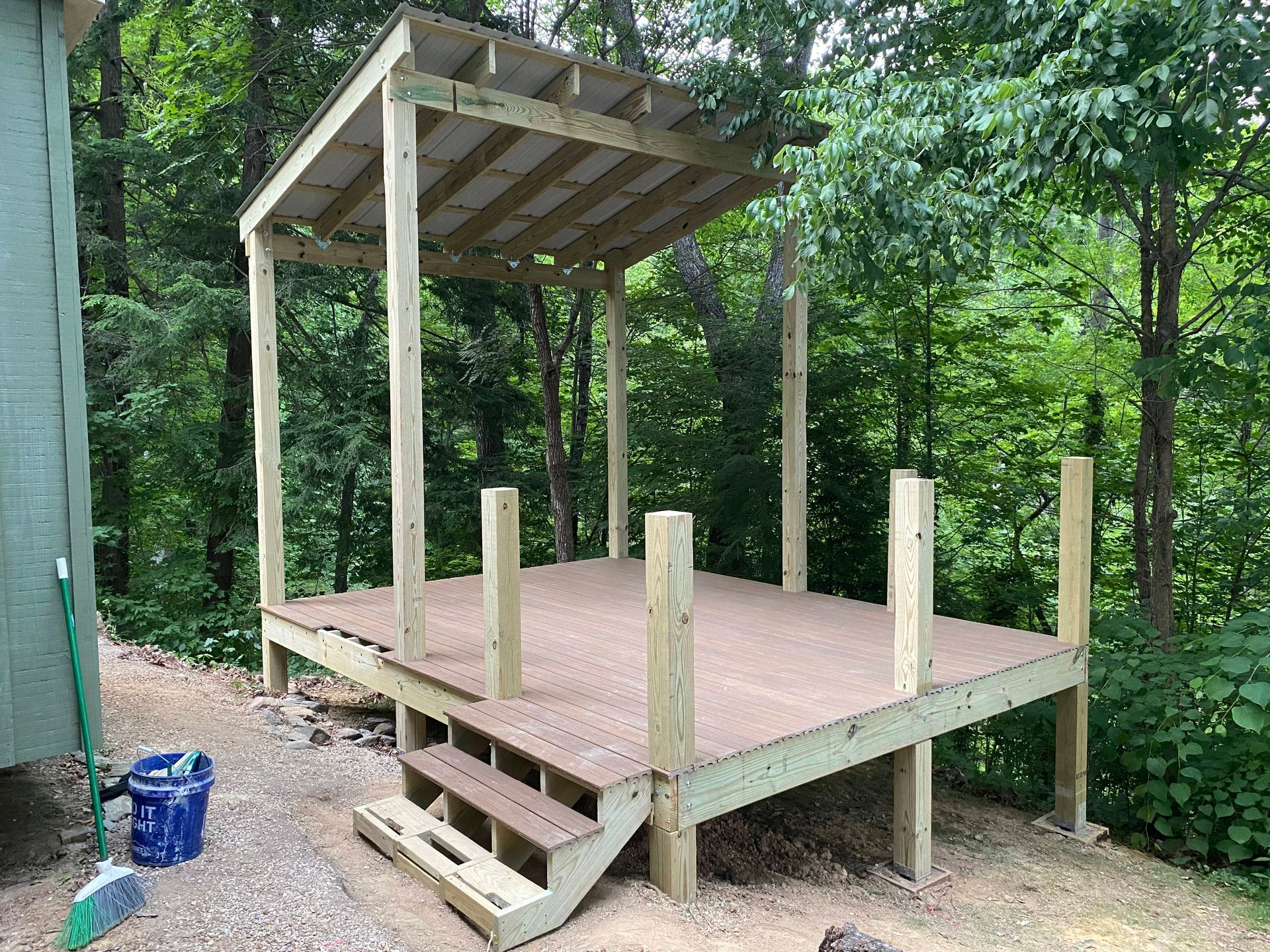 Wooden deck with steps and a small roofed structure in a wooded area.