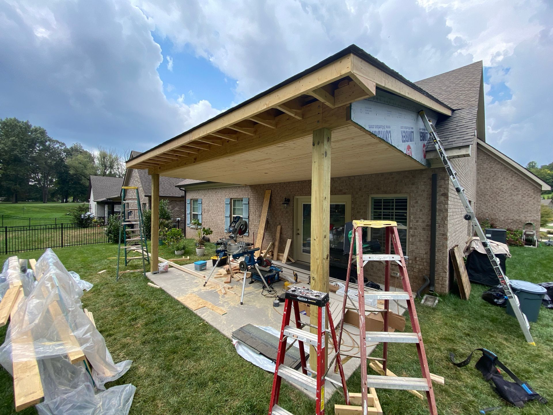Construction of a covered patio, with a ladder leaning against the brick house, tools scattered on the patio.