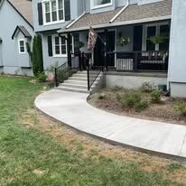 A concrete walkway leads to a house with a black front door. Lush green grass surrounds it.