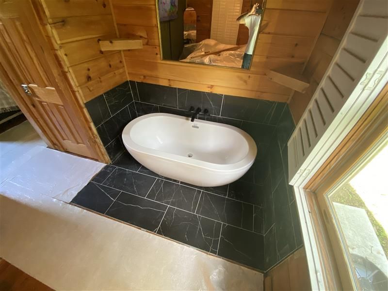 Bathroom with oval tub, black tiled floor, and wood paneling. Sunlight streams in from a window.