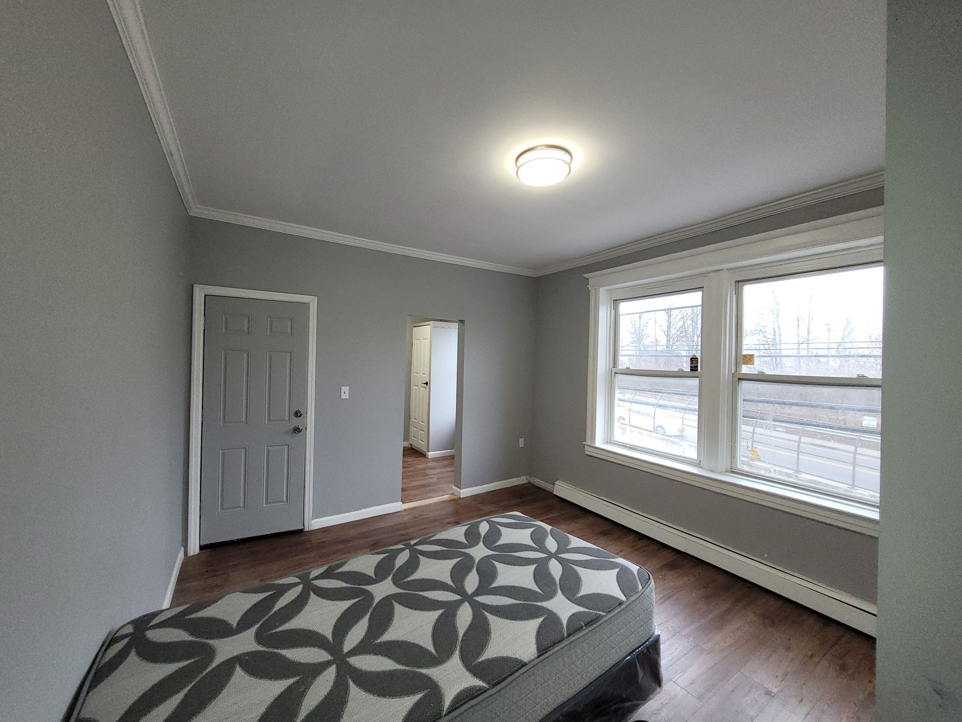 Bedroom with gray walls, white trim, hardwood floor, bed with patterned comforter, window.