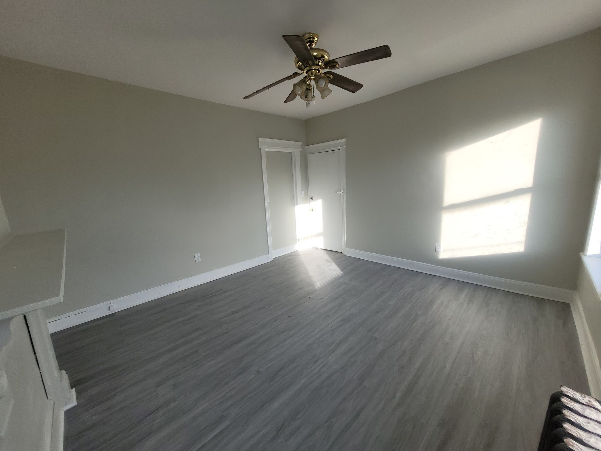 Empty room with gray walls, flooring, and ceiling fan. Sunlight through window illuminates space.