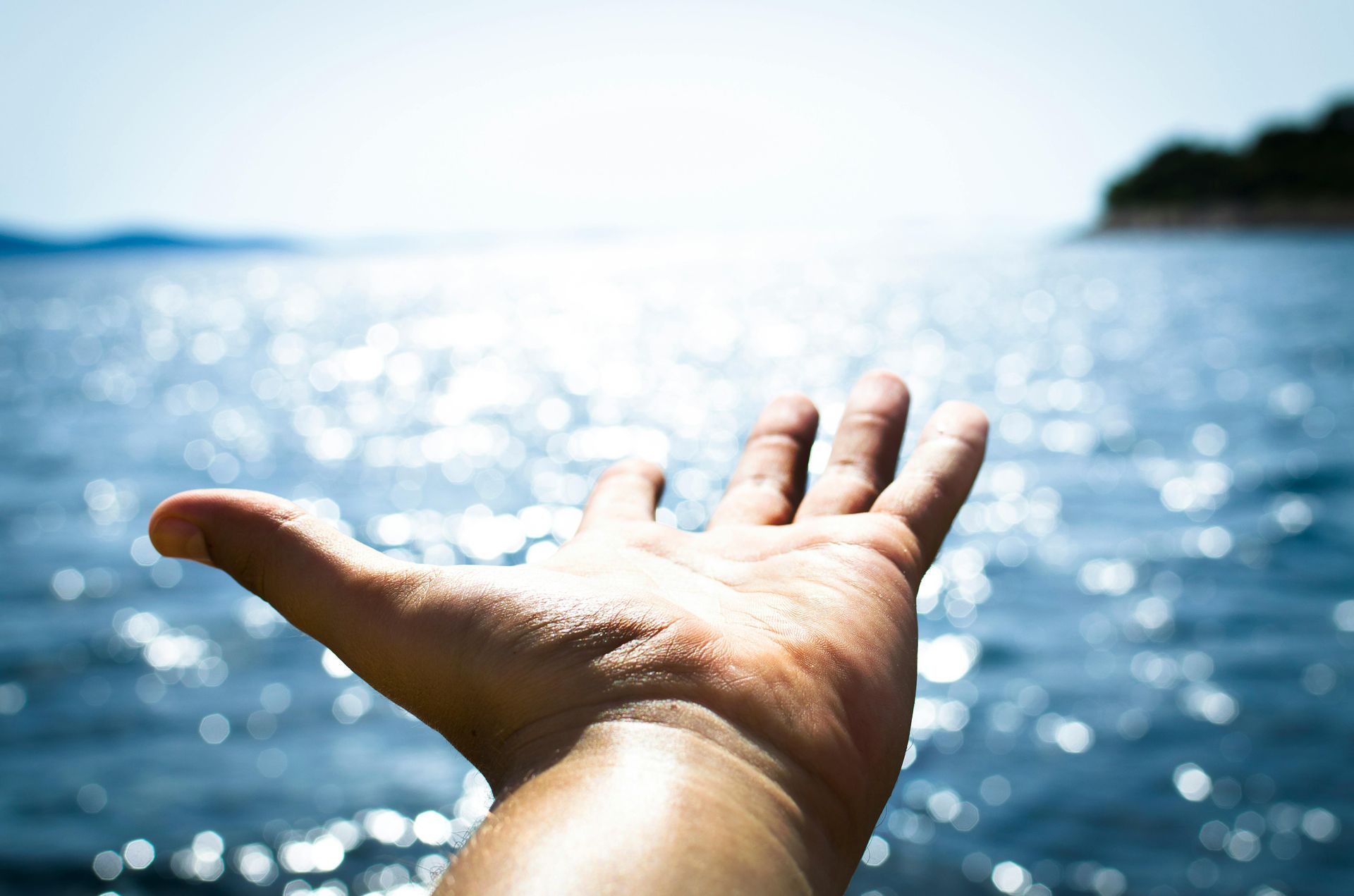 Hand reaching toward shimmering blue water and bright sky.