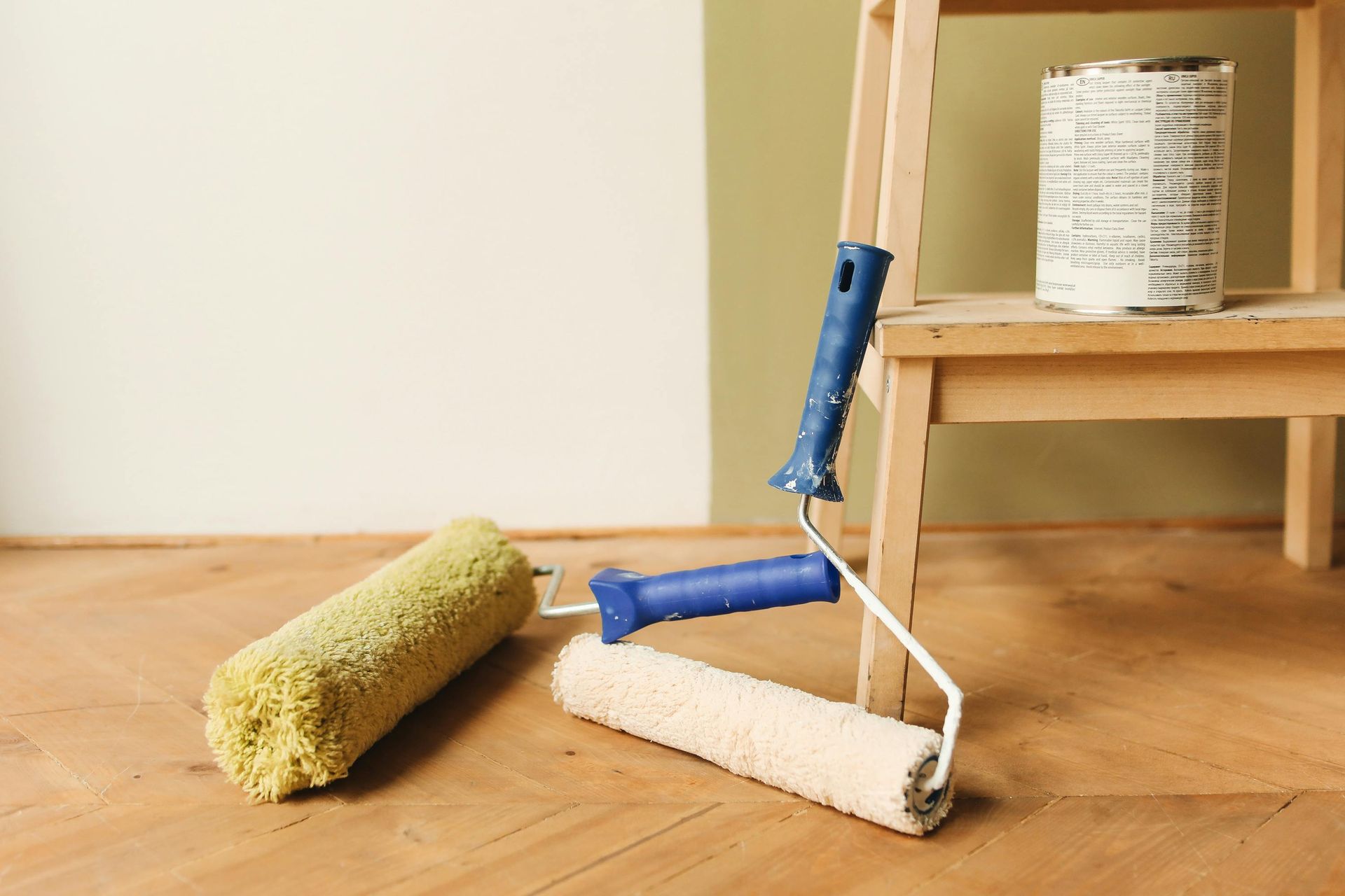 Paint rollers and a can of paint near a step ladder on a wooden floor in front of two-toned walls.