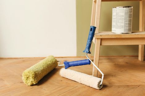 Paint rollers and a can of paint near a step ladder on a wooden floor in front of two-toned walls.