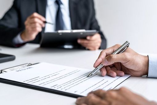 Person signing a document on a clipboard during a meeting. Another person is in the background.