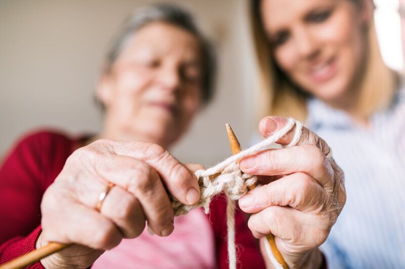 A care giver is helping an elderly woman knit.