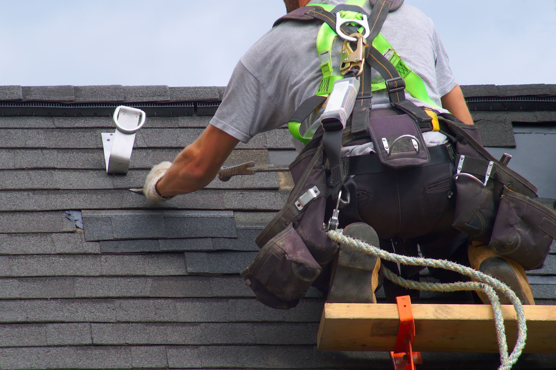 A roofer wearing a secured rope is performing roof repair on a house.
