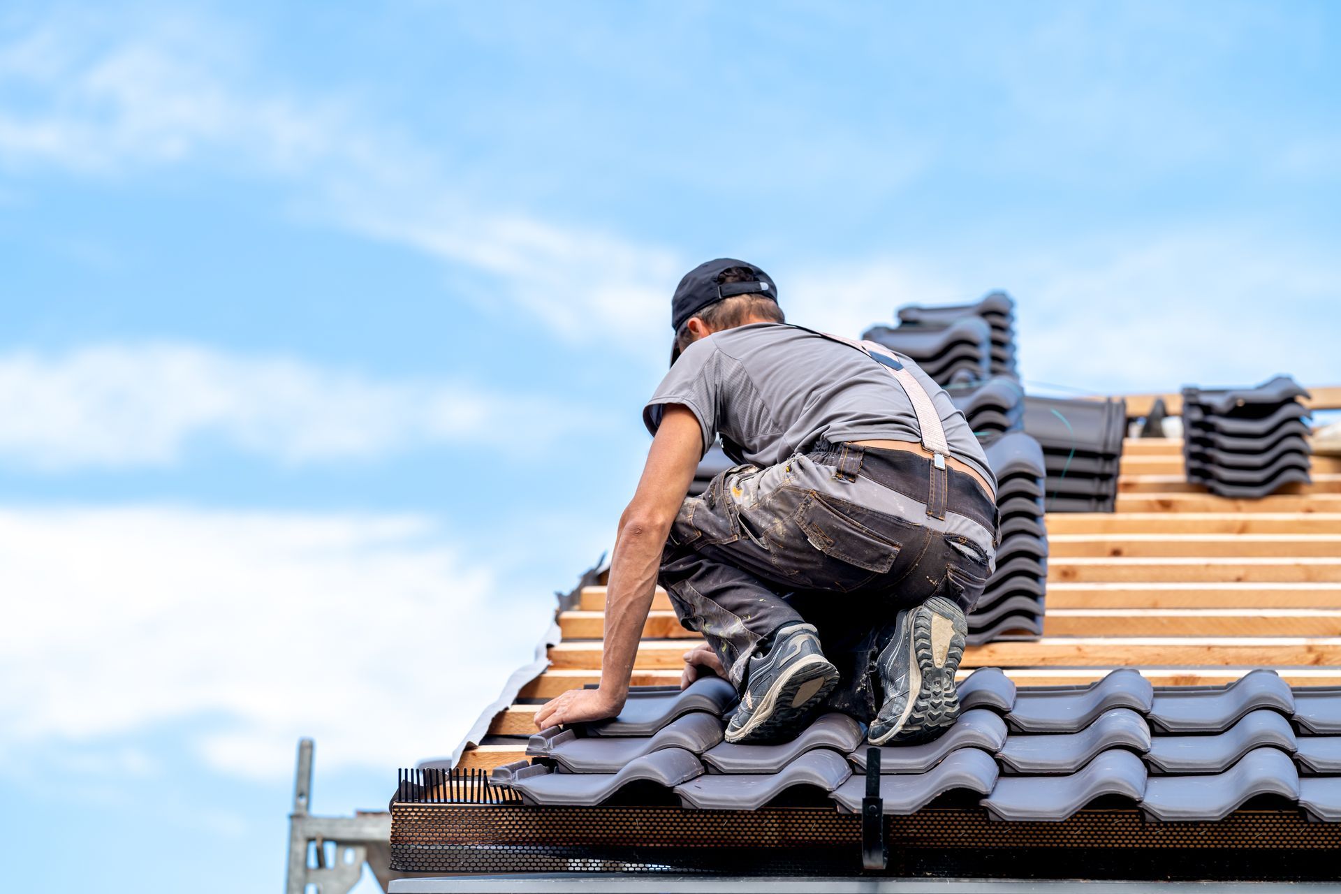 A roofer crouches on the roof, fitting dark tiles beneath a clear sky. New roof installation