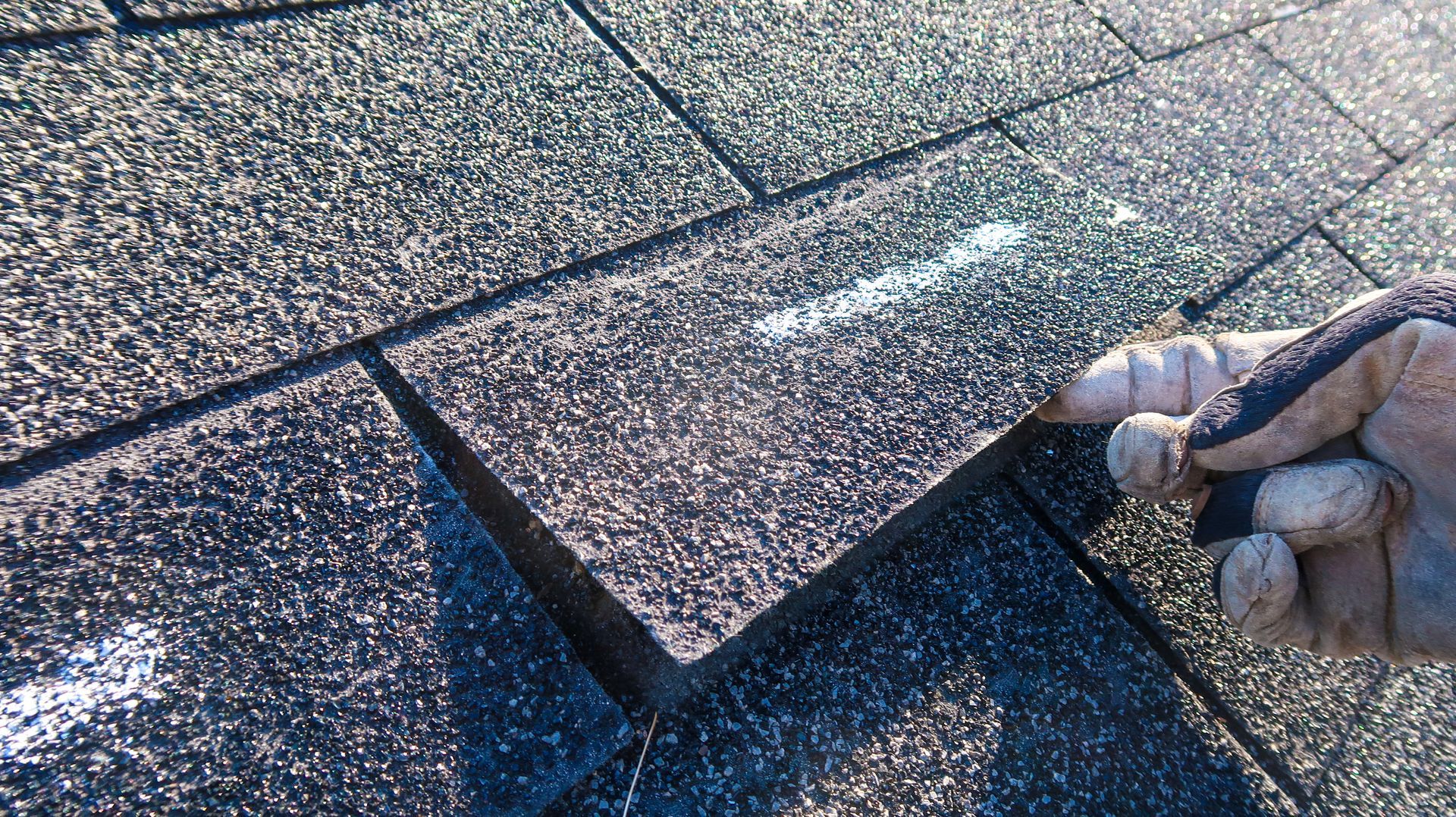 A gloved hand lifts the edge of a dark asphalt roof shingle, revealing a chalk mark on the surface beneath.