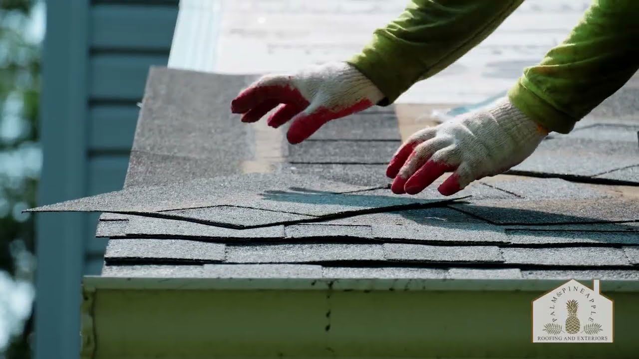 Hands wearing gloves placing roofing shingles on a rooftop; outdoor setting.