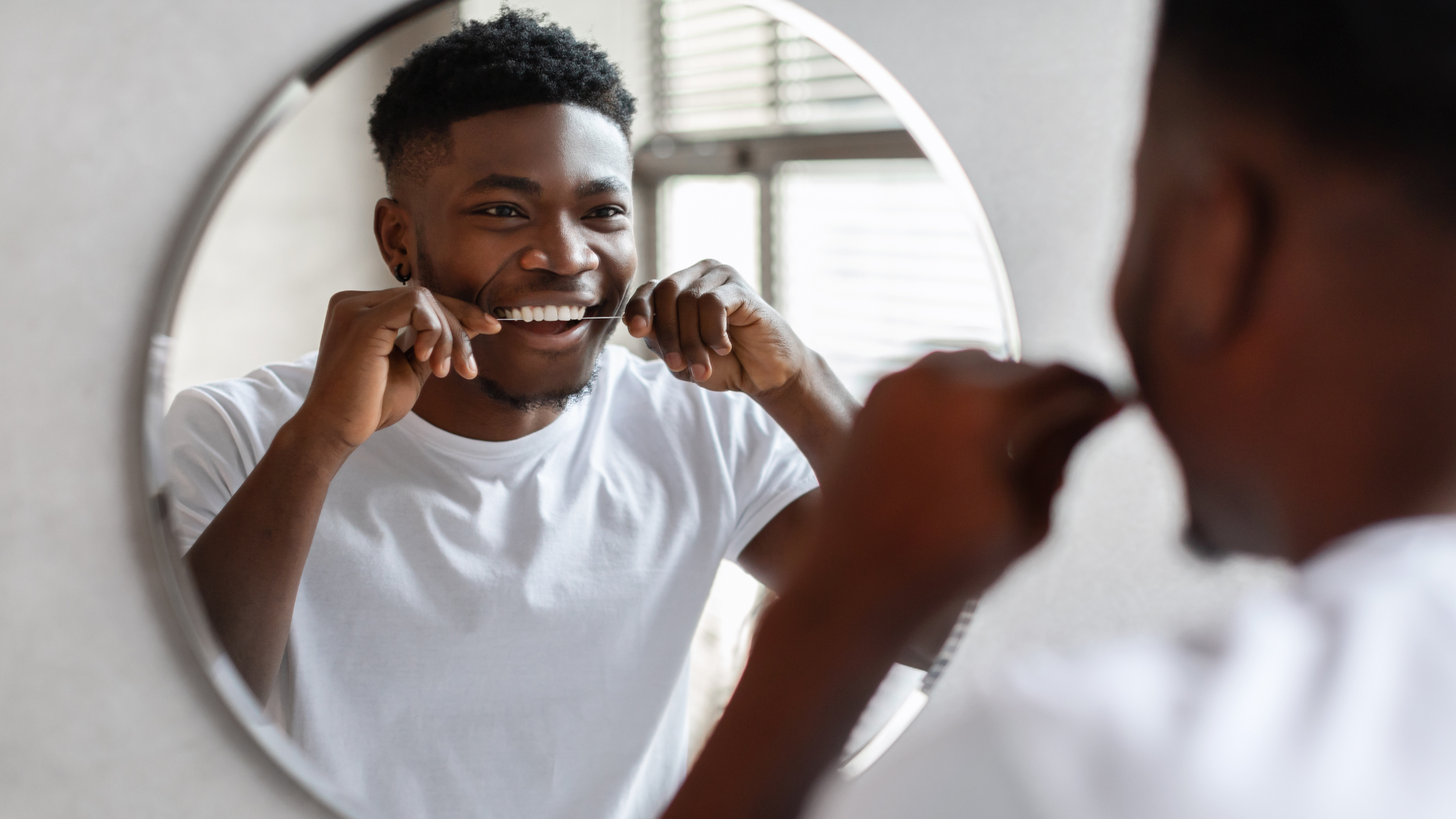 Man flossing his teeth in the mirror; wearing a white shirt, smiling.
