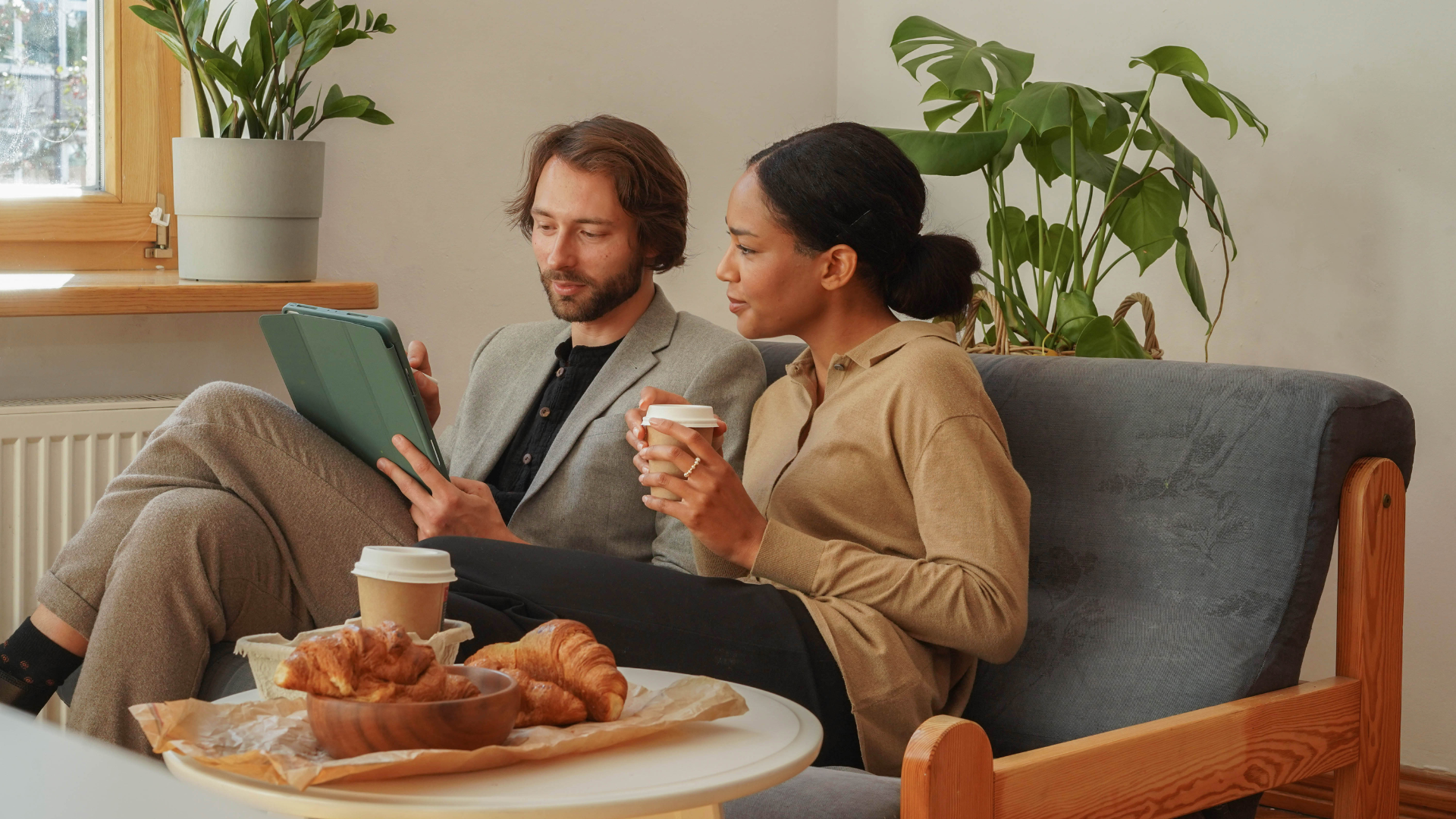 A man and a woman are sitting on a couch reading a book and drinking coffee.