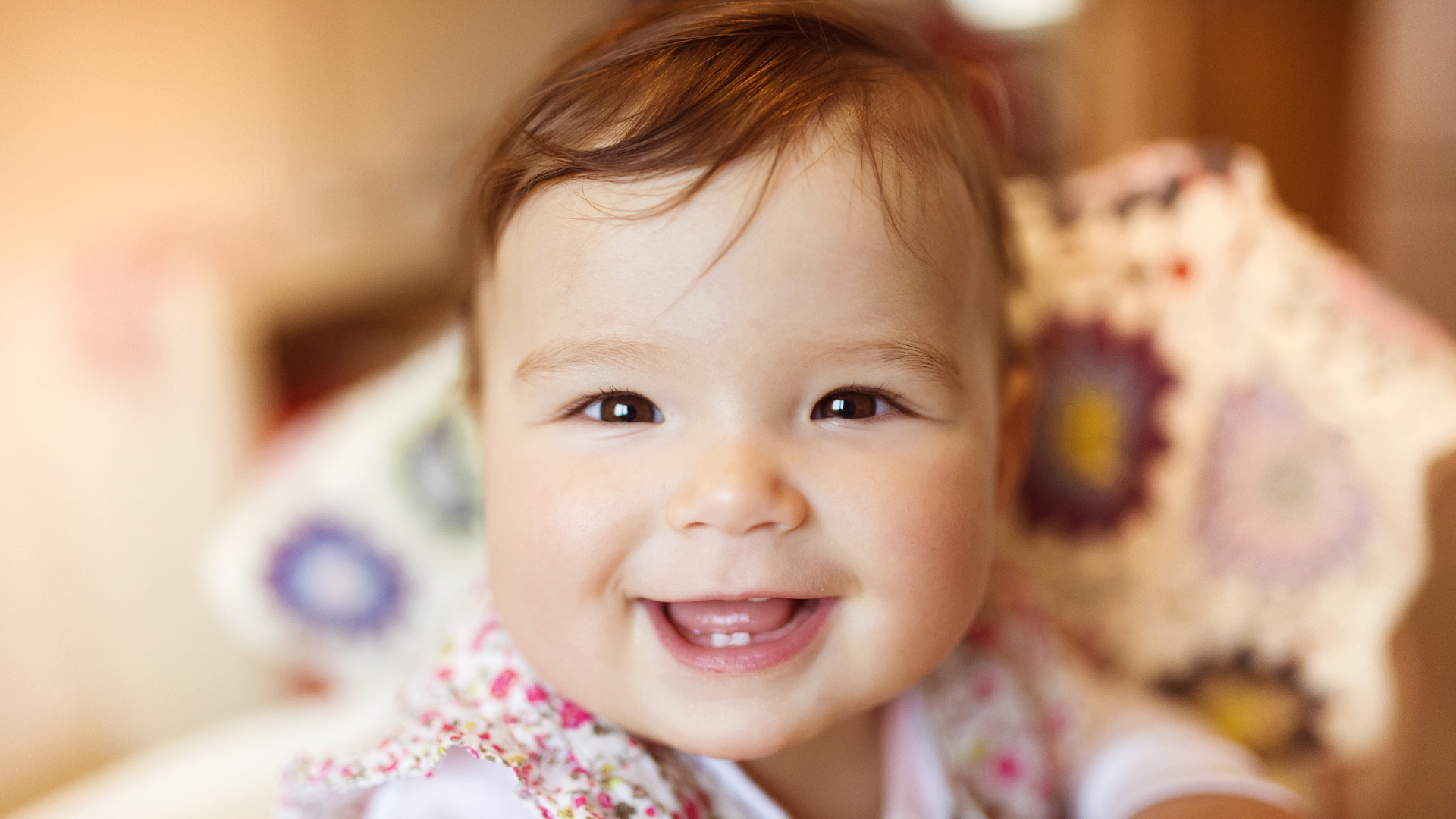 Smiling baby with brown hair and eyes, wearing a floral bib, sitting in a high chair.