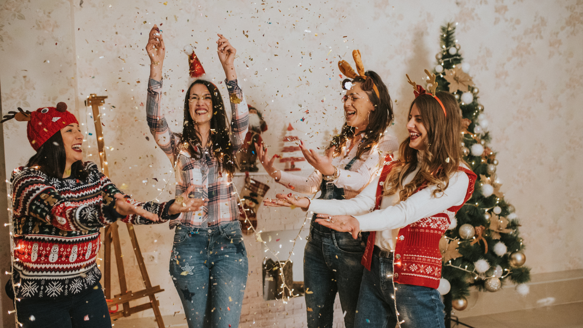Four women celebrating, throwing confetti near a Christmas tree; festive sweaters, antlers, and happy expressions.