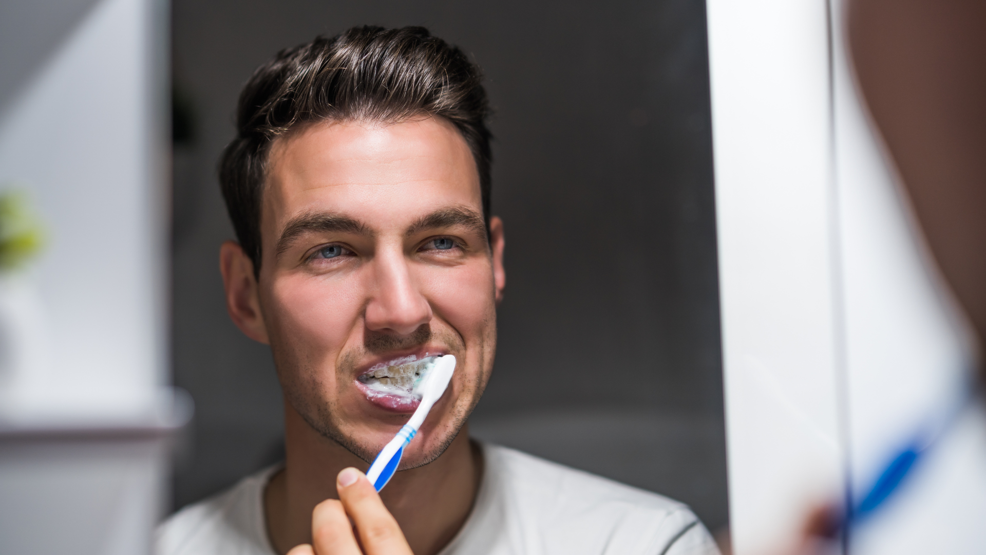 Man brushing his teeth in bathroom, looking in mirror.