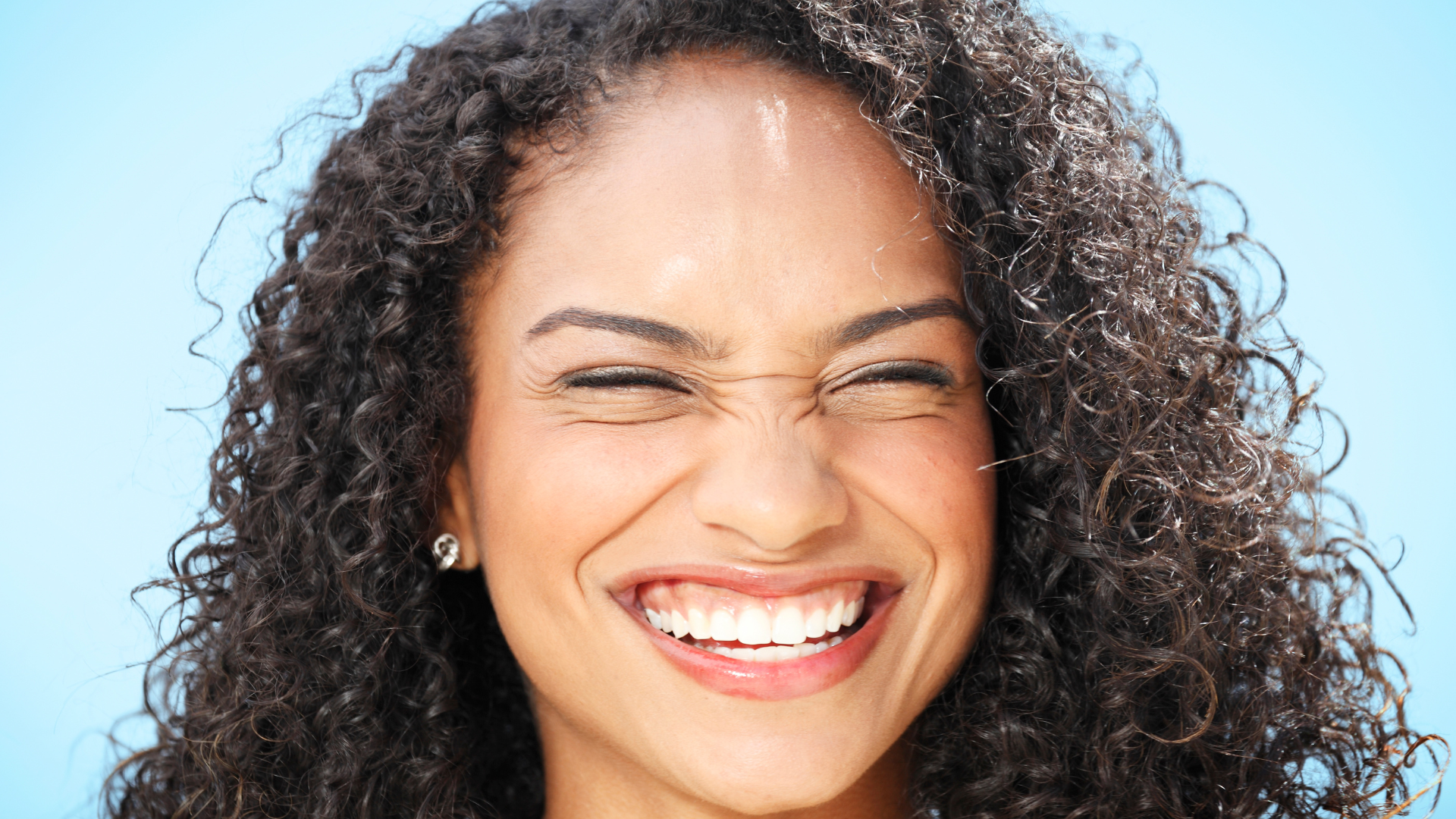 Woman with curly hair smiles broadly, eyes closed against a light blue background.