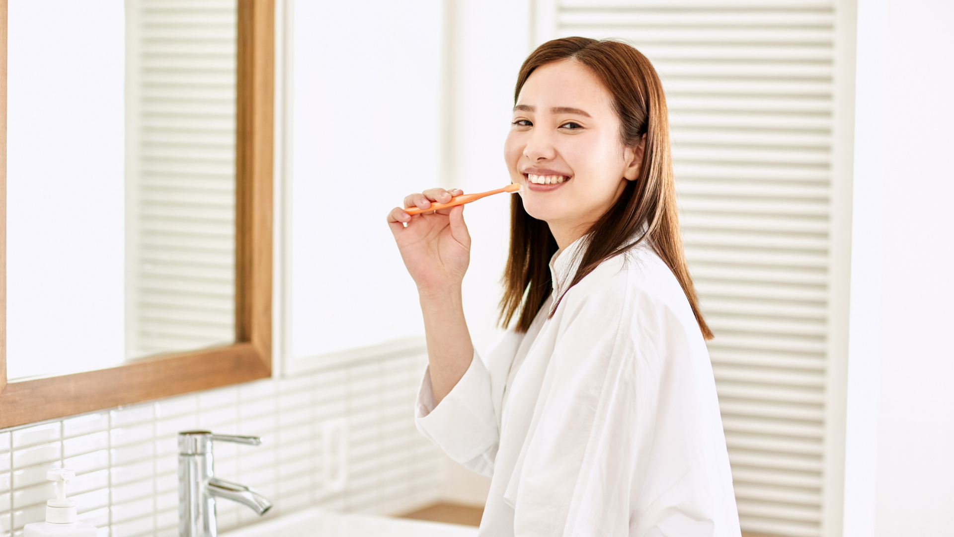 Woman brushing teeth in bathroom, smiling, wearing a white robe.