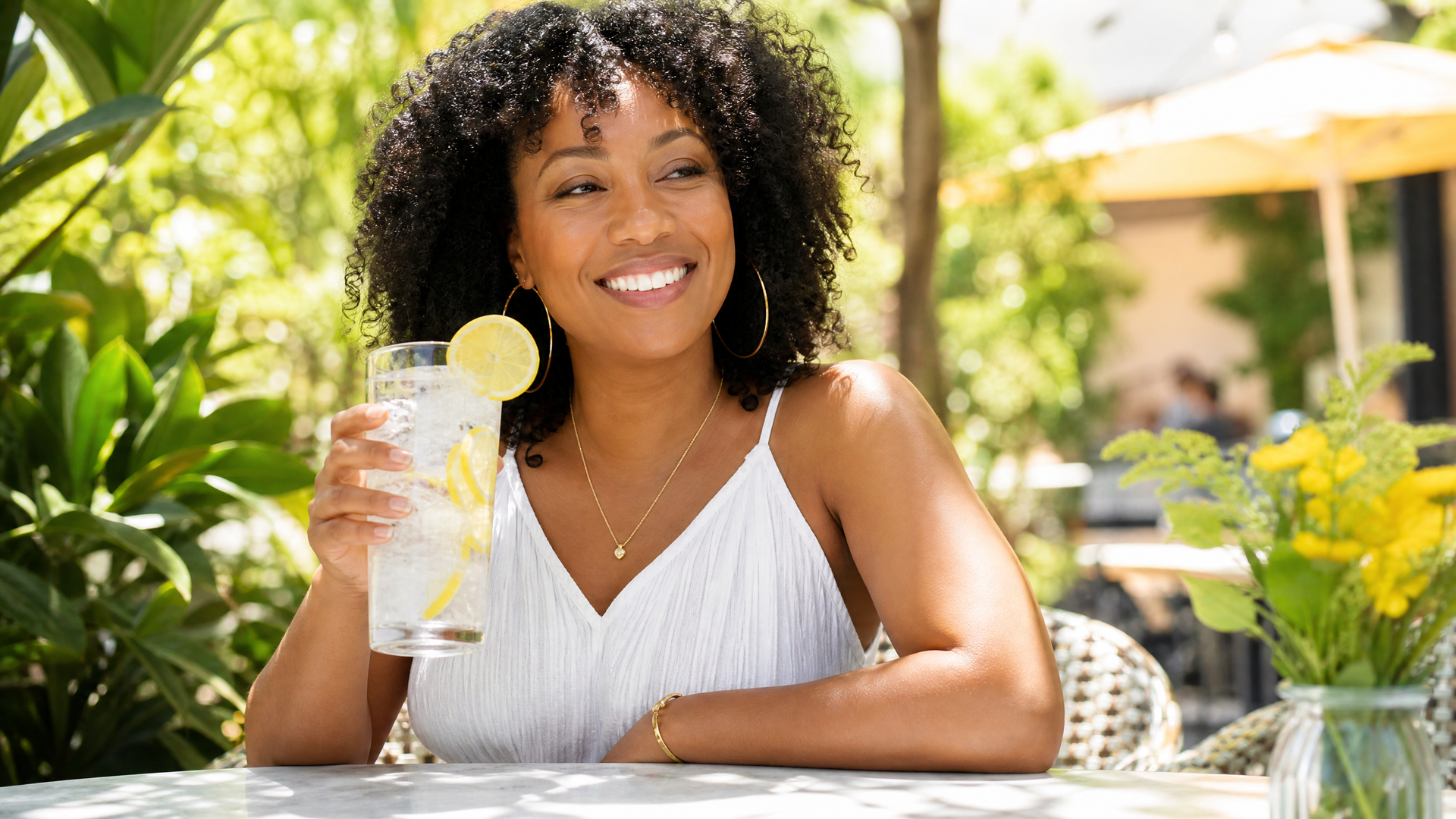 Smiling woman holding a lemon drink at an outdoor table in a sunny garden.