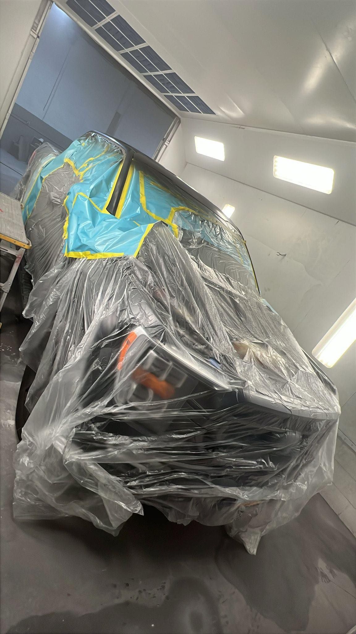A car partially covered in protective plastic wrap inside an auto body paint booth.