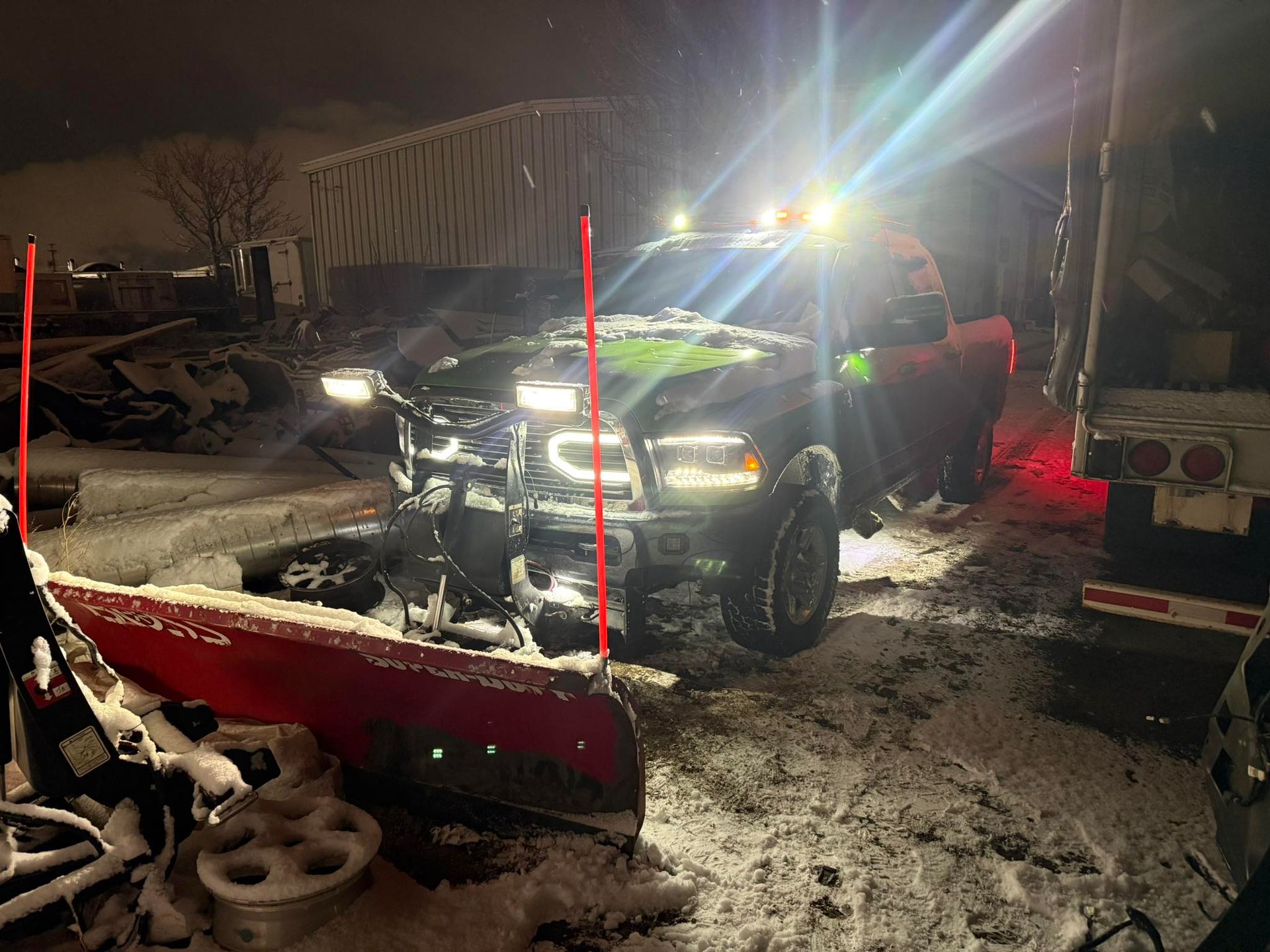 A dark pickup truck equipped with a large red snowplow, parked in a snowy area at night with its lights on.