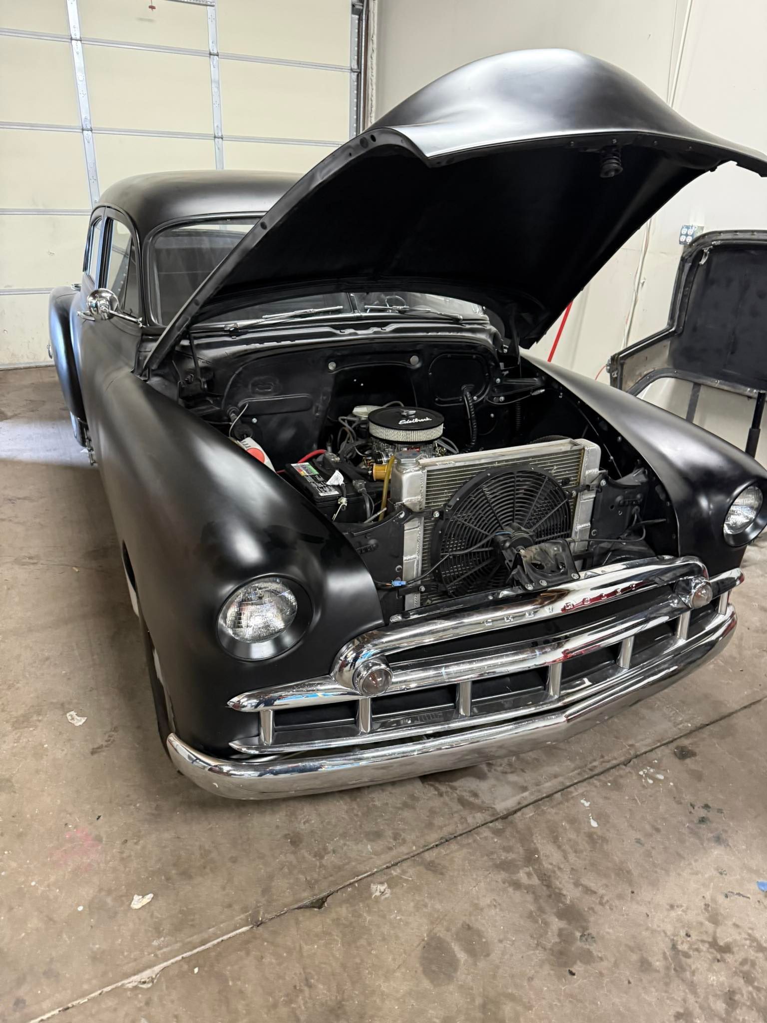 A matte black vintage car parked in a garage with its hood open, revealing the engine.