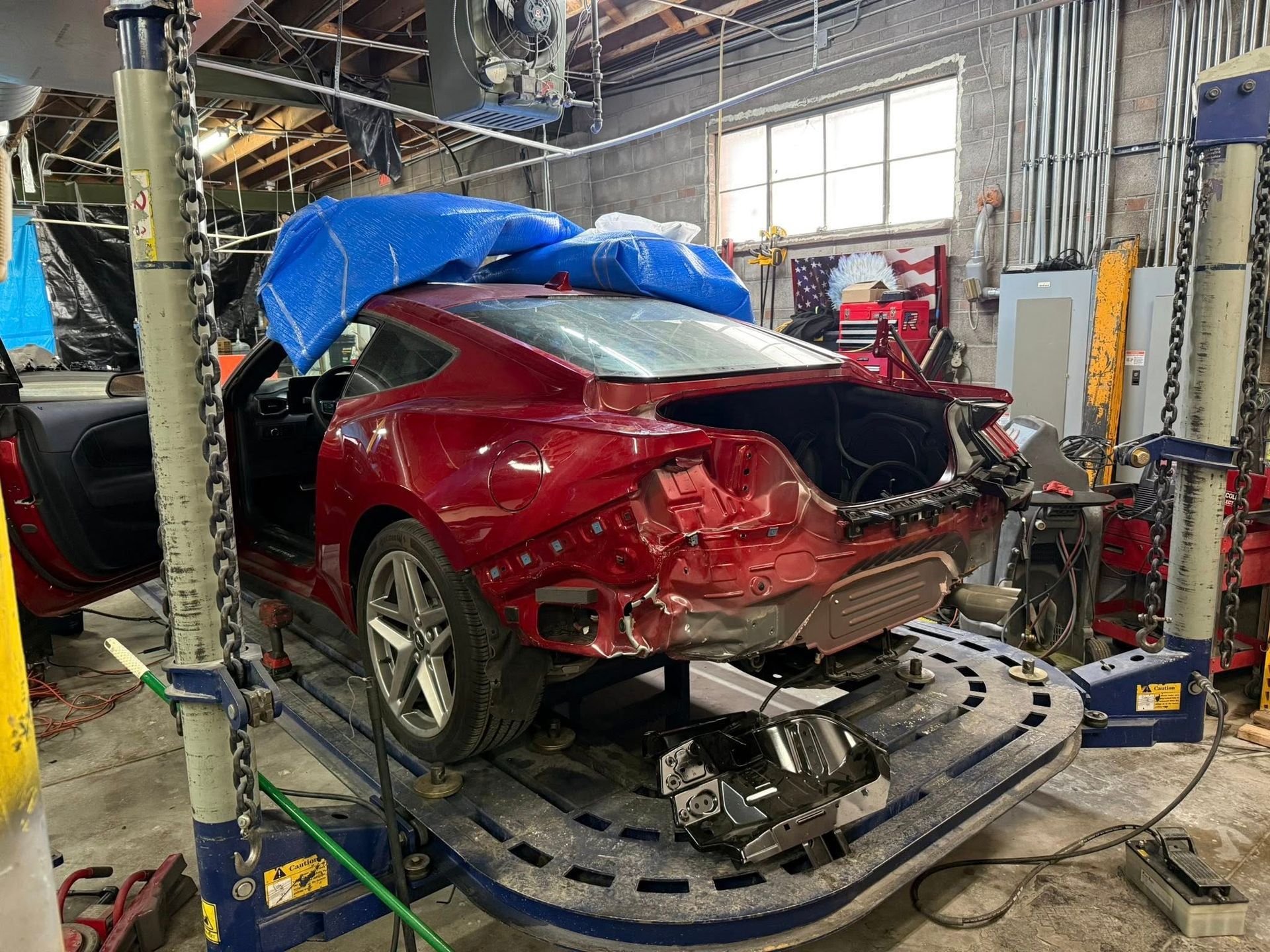 A red sports car with a removed rear bumper sits on a structural frame machine inside an auto body repair shop.