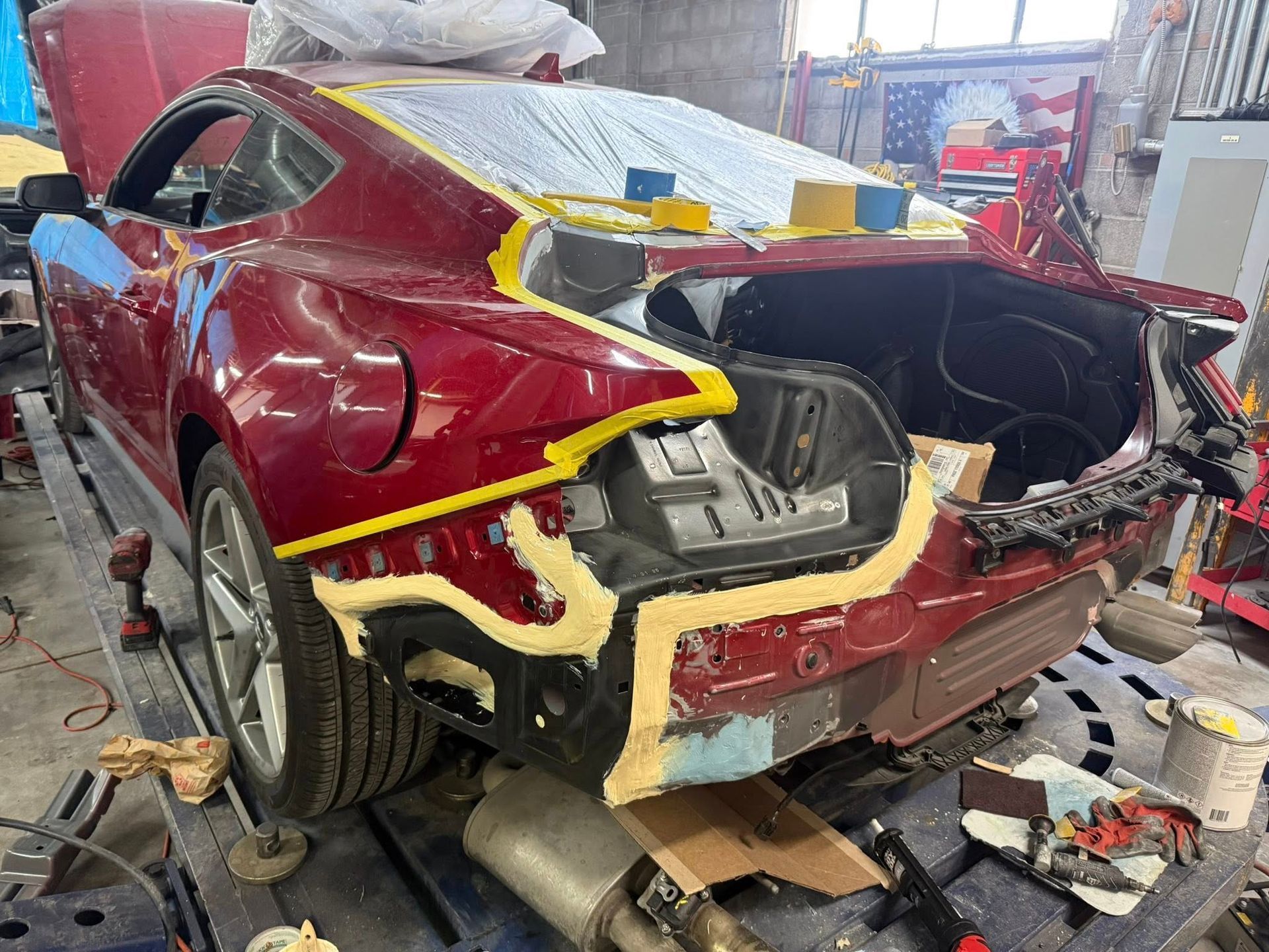 A red car undergoing body repair in a workshop, featuring yellow masking tape and exposed panels at the rear.