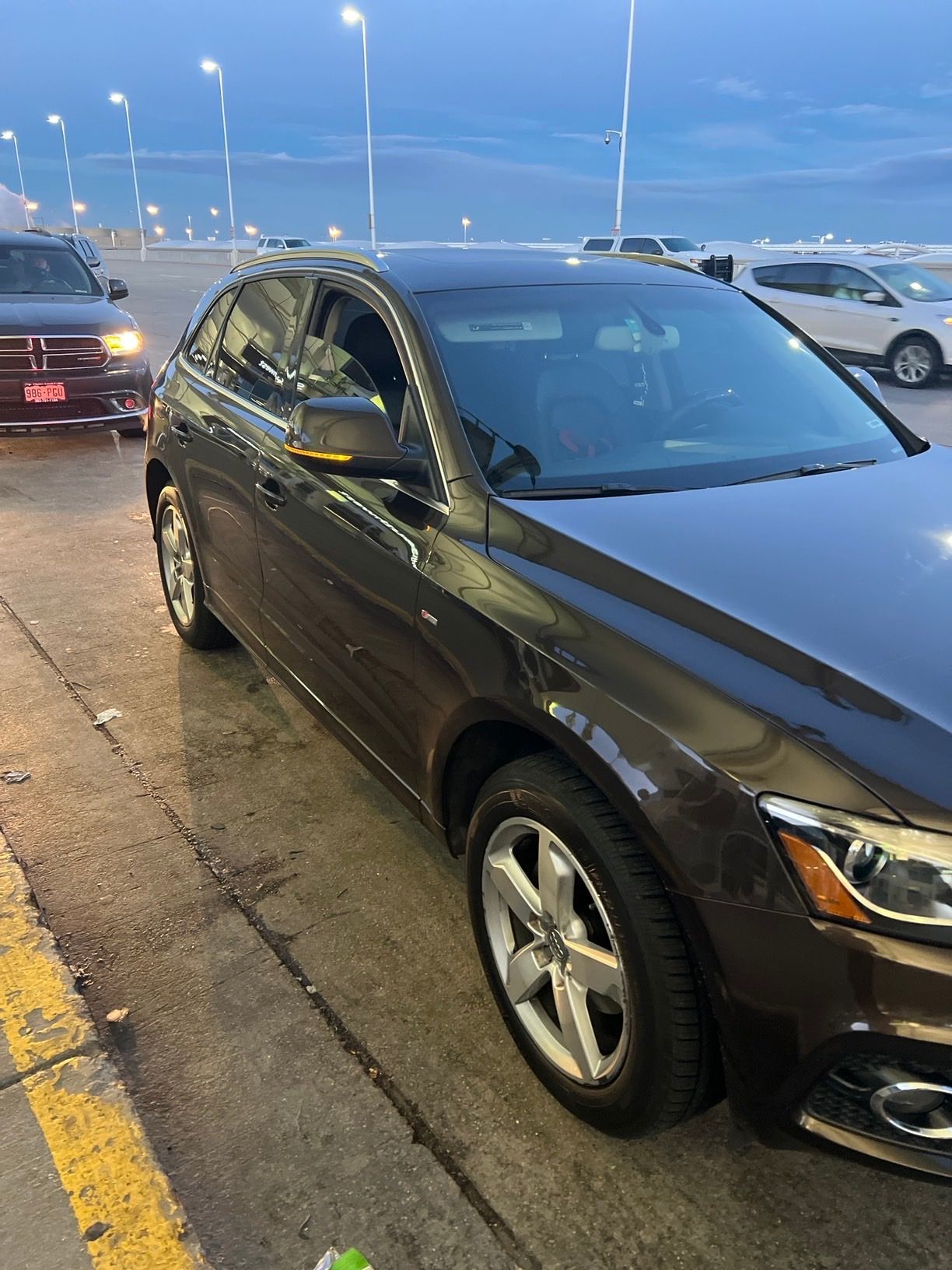 A dark-colored sedan parked in a lot during twilight, with other vehicles visible nearby under outdoor lighting.