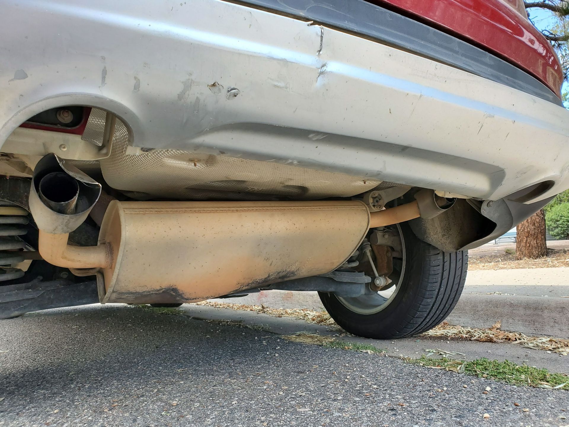 The rear underside of a car showing the muffler, exhaust pipe, and rear bumper, parked on an asphalt road.