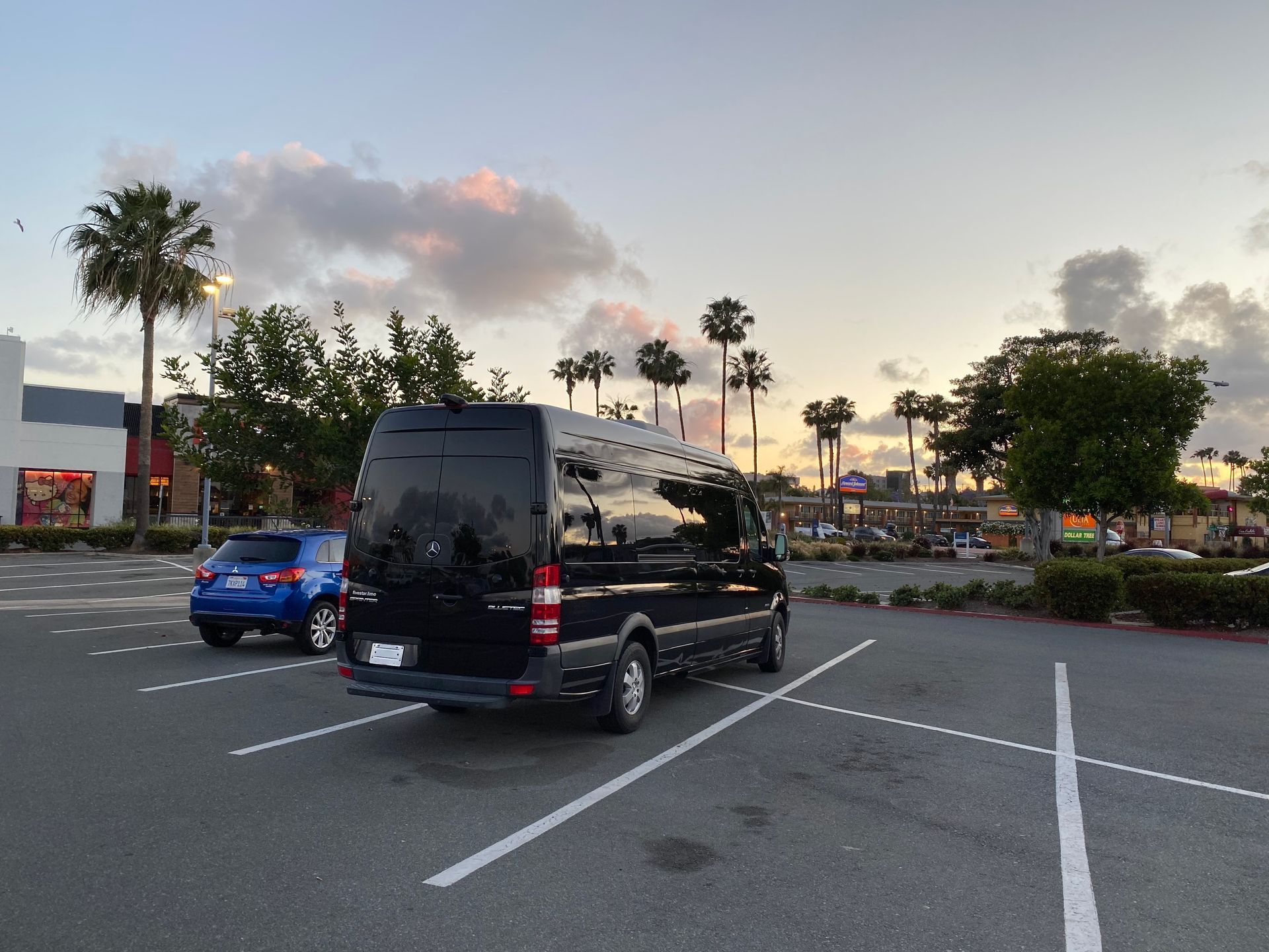 A black passenger van and a blue car parked in an outdoor shopping center parking lot during sunset.