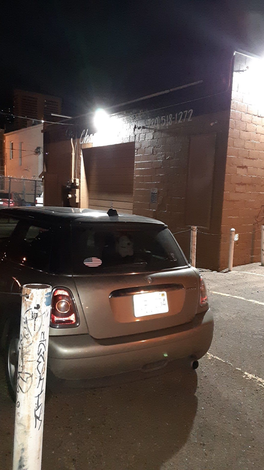 A silver Mini Cooper parked in a dimly lit outdoor lot at night, in front of a dark brick building with an open garage bay.
