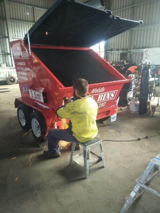 A Man is Sitting on a Stool in Front of a Dumpster — Hastings Scrap Metal Recyclers in Kundabung, NSW