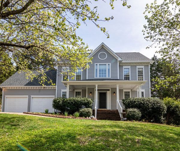 A large house with a large porch and two garages is sitting on top of a lush green hill.