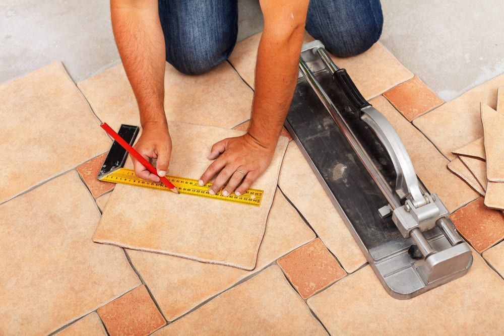 Person on Knees Measuring a Tile With a Ruler and Pencil, With a Tile Cutter Nearby — Cooloola Tile Company in Gympie, QLD