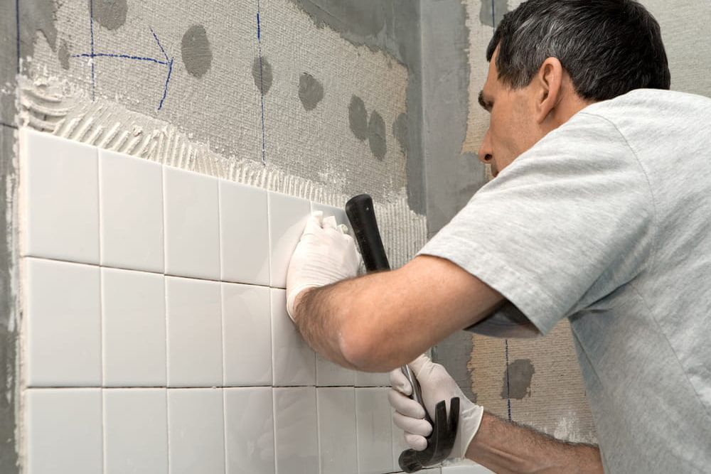 Man Tiling a Wall With White Tiles, Wearing Gloves, Using a Hammer — Cooloola Tile Company in Gympie, QLD