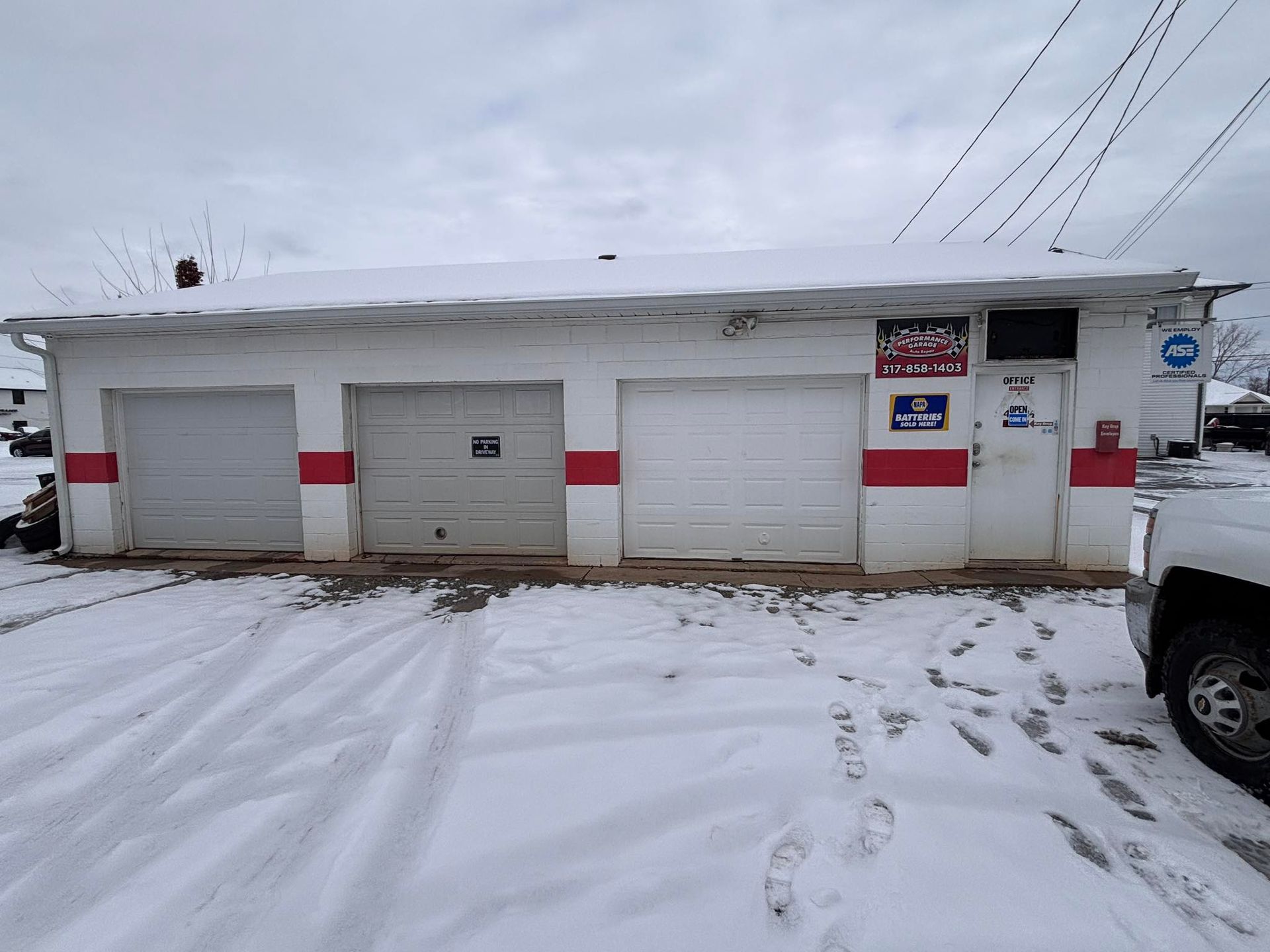 A white, one-story garage building with three bay doors, a red horizontal stripe, and a snow-covered exterior.