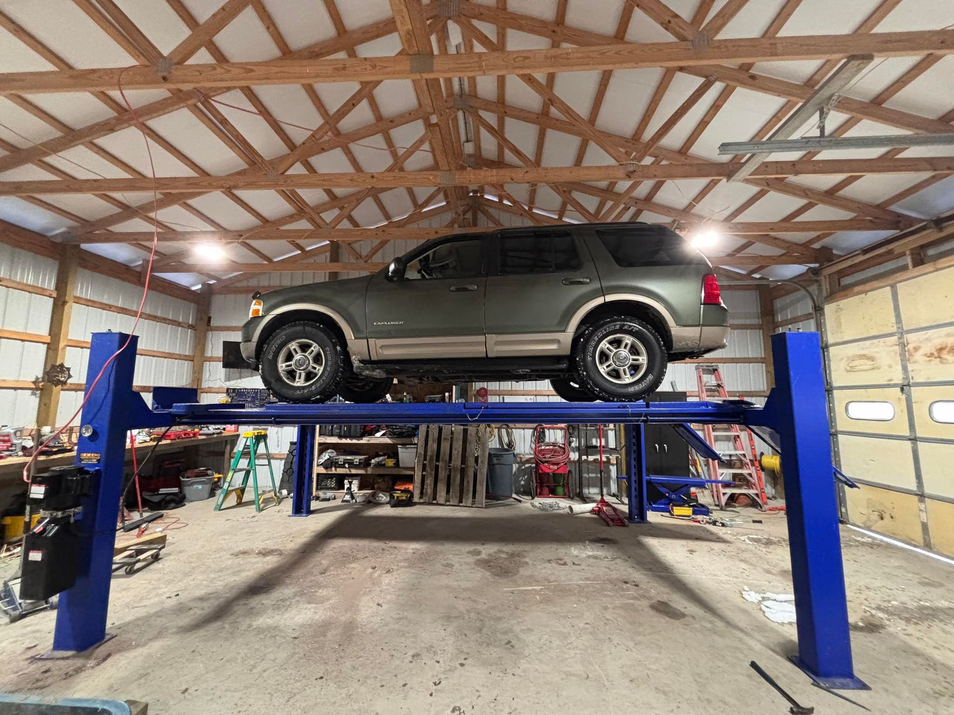 A dark green SUV raised on a blue four-post automotive lift inside a workshop with a wooden-truss ceiling.