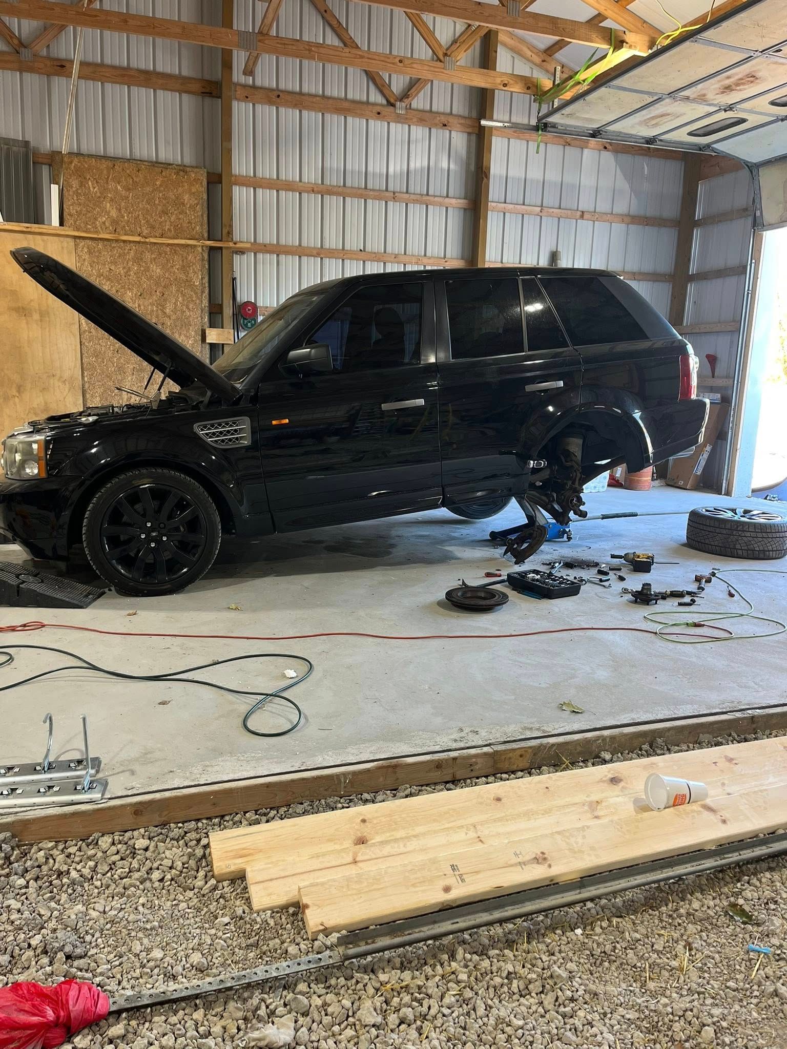 A black SUV with its hood up and rear wheel removed, undergoing repairs in a workshop with concrete floors.