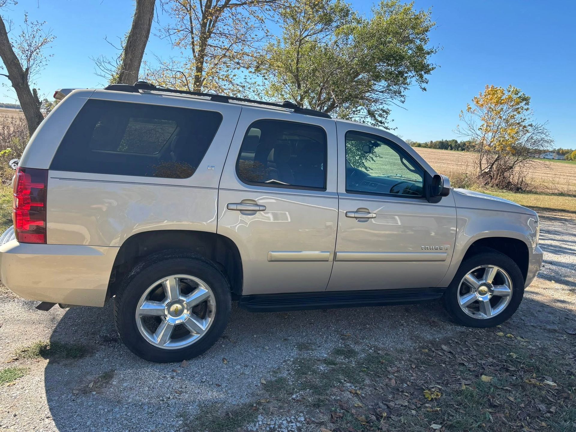 A gold Chevrolet Tahoe SUV parked on a gravel lot in a rural setting with trees in the background.