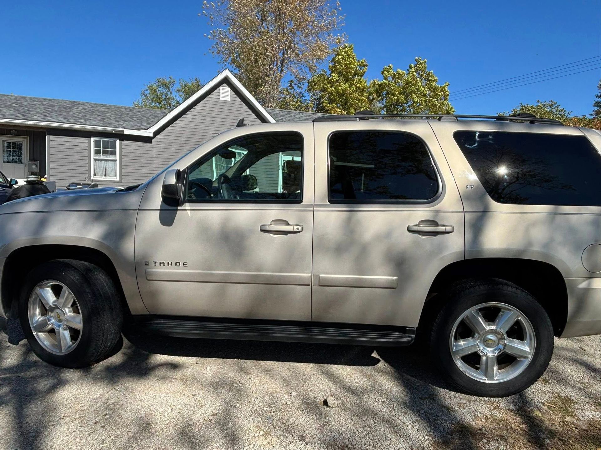 A tan Chevrolet Tahoe SUV parked on a gravel driveway in front of a house on a sunny day.