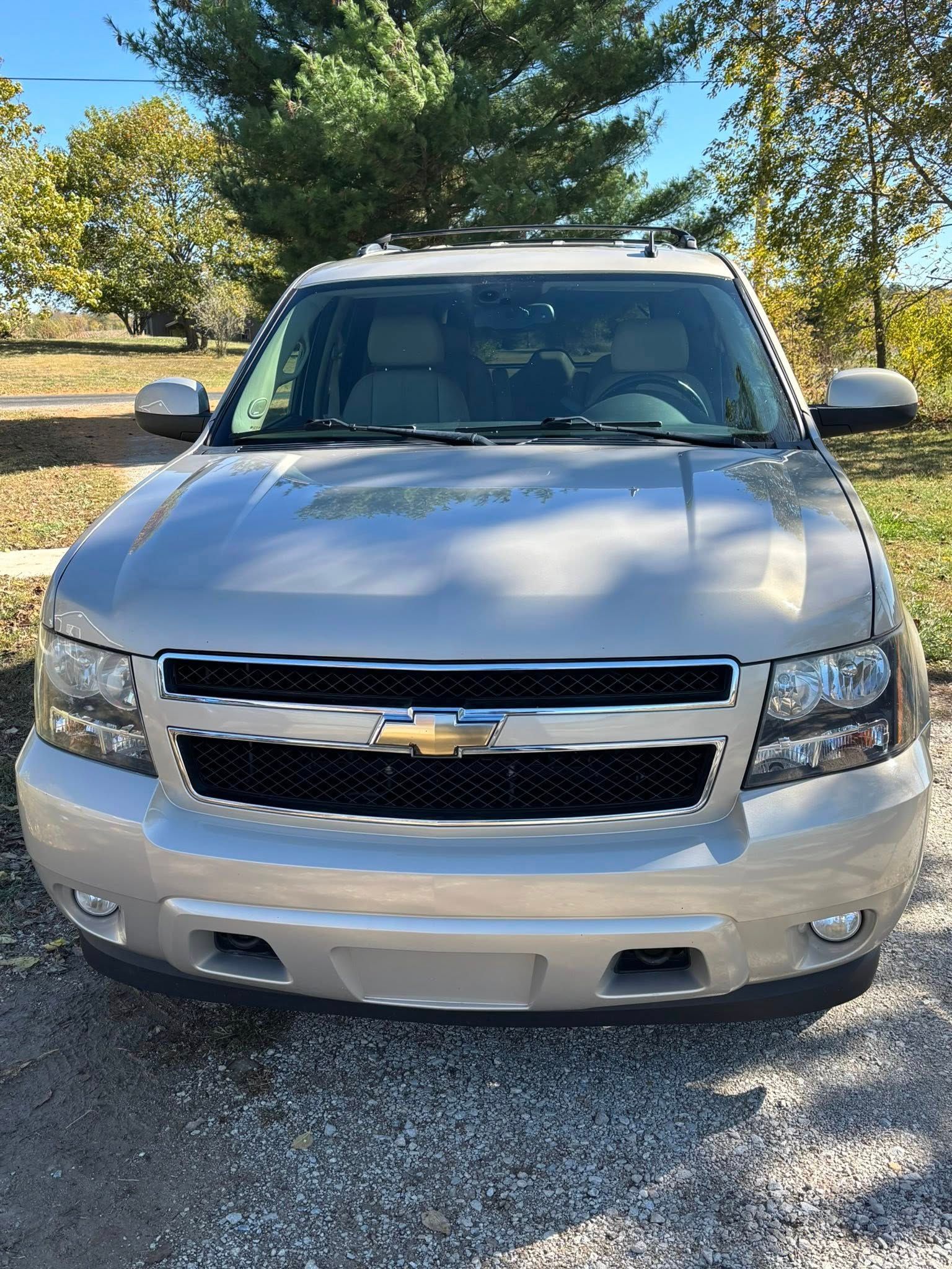 A silver Chevrolet Tahoe SUV parked on a gravel surface with trees in the background.