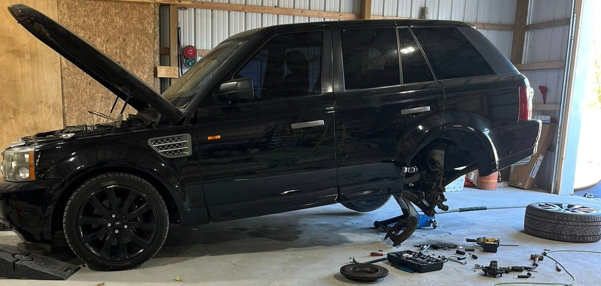 A black SUV in a garage with its hood open, rear wheel removed, and disassembled brake parts on the concrete floor.