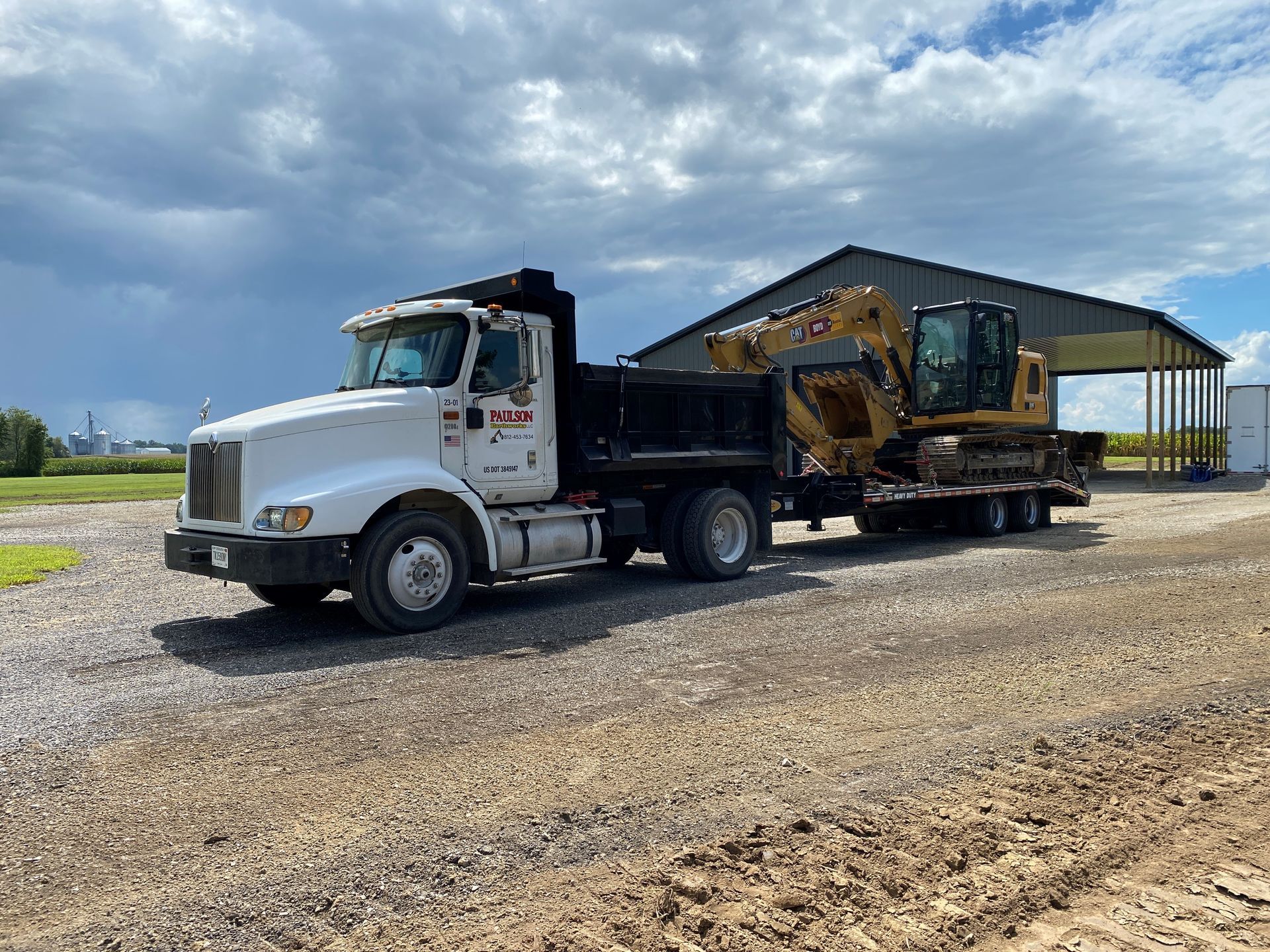 A white dump truck is carrying a yellow excavator on a trailer.