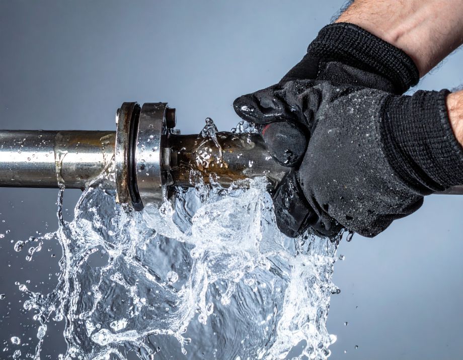 Hands wearing black gloves gripping a leaking metal pipe, water spraying outwards.